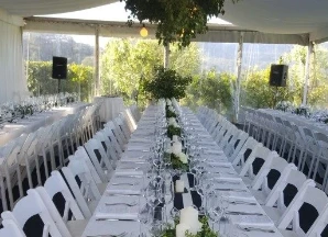 Marquee with white roof and clear walls for event furnished with white chairs and linen. Decorated with a beautiful green pendant and other decor.